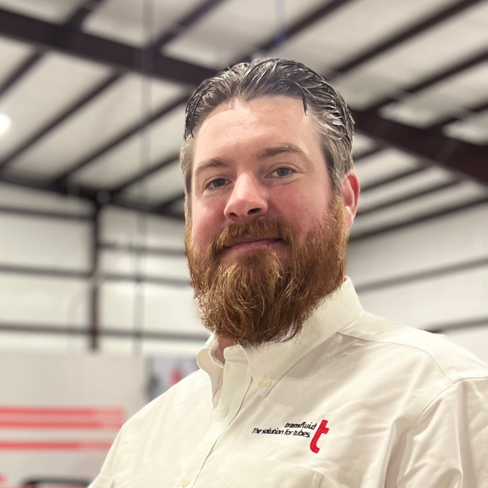 Porträt eines lächelnden Mitarbeiters mit Bart in einer Produktionshalle, getragenes weißes Hemd mit Firmenlogo.