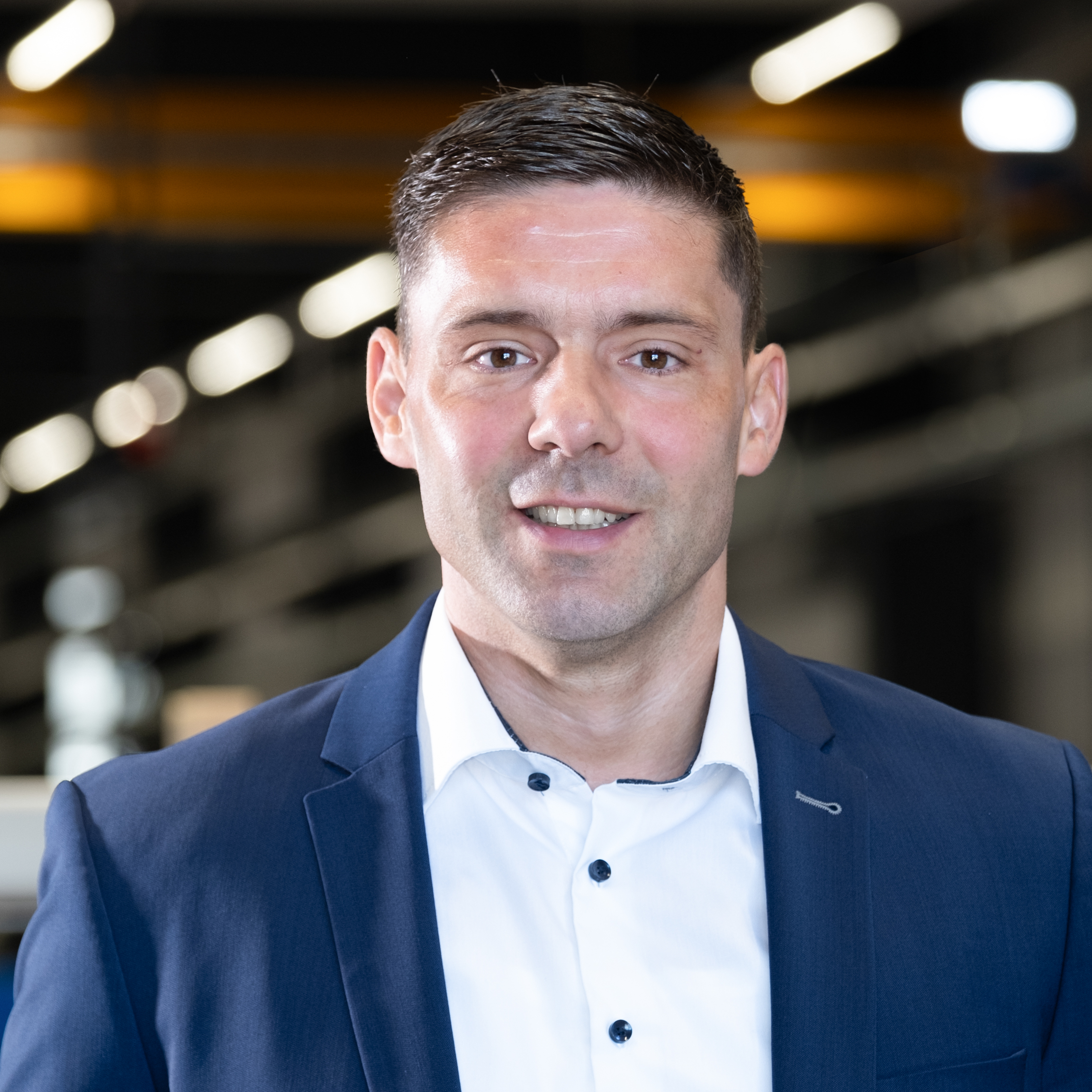 Mitarbeiter im blauen Anzug in moderner Industriehalle mit unscharfem Maschinenhintergrund.