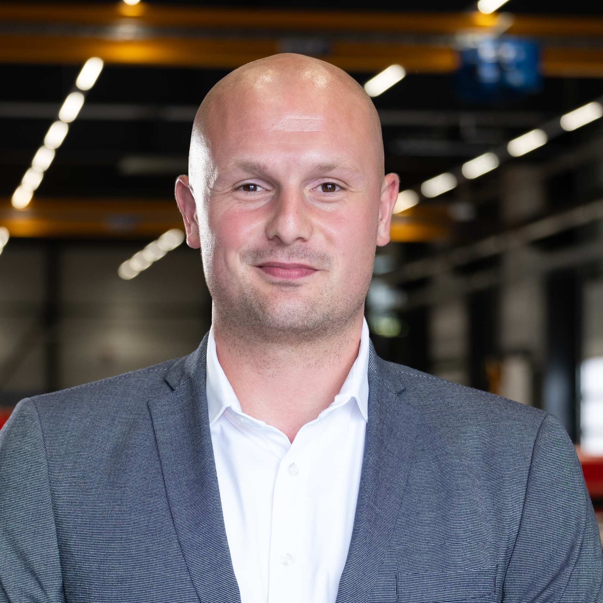 Mitarbeiter im grauen Sakko in Industriehalle mit unscharfem Maschinenhintergrund.