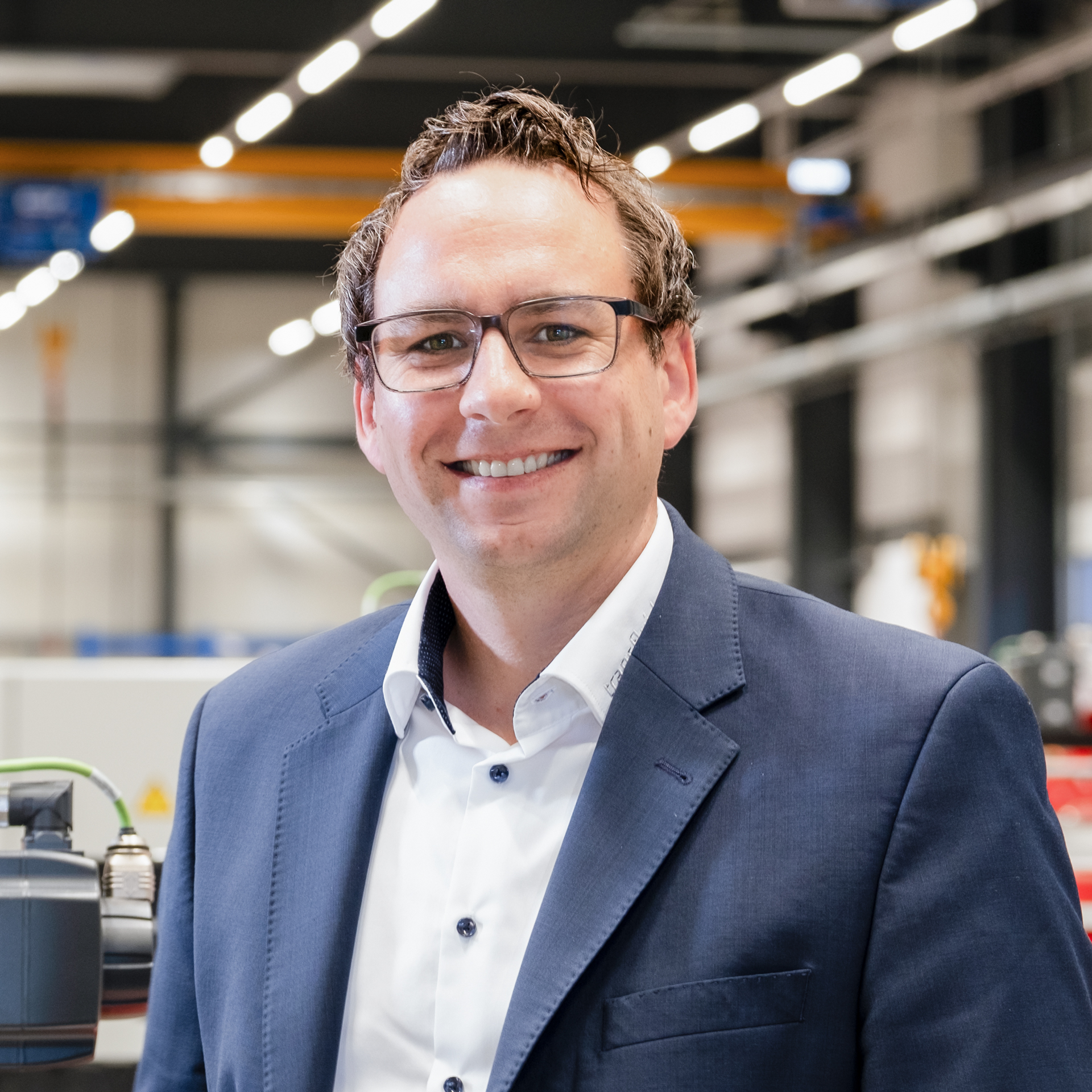 Mitarbeiter mit Brille im blauen Anzug in moderner Industriehalle mit Maschinen im Hintergrund.