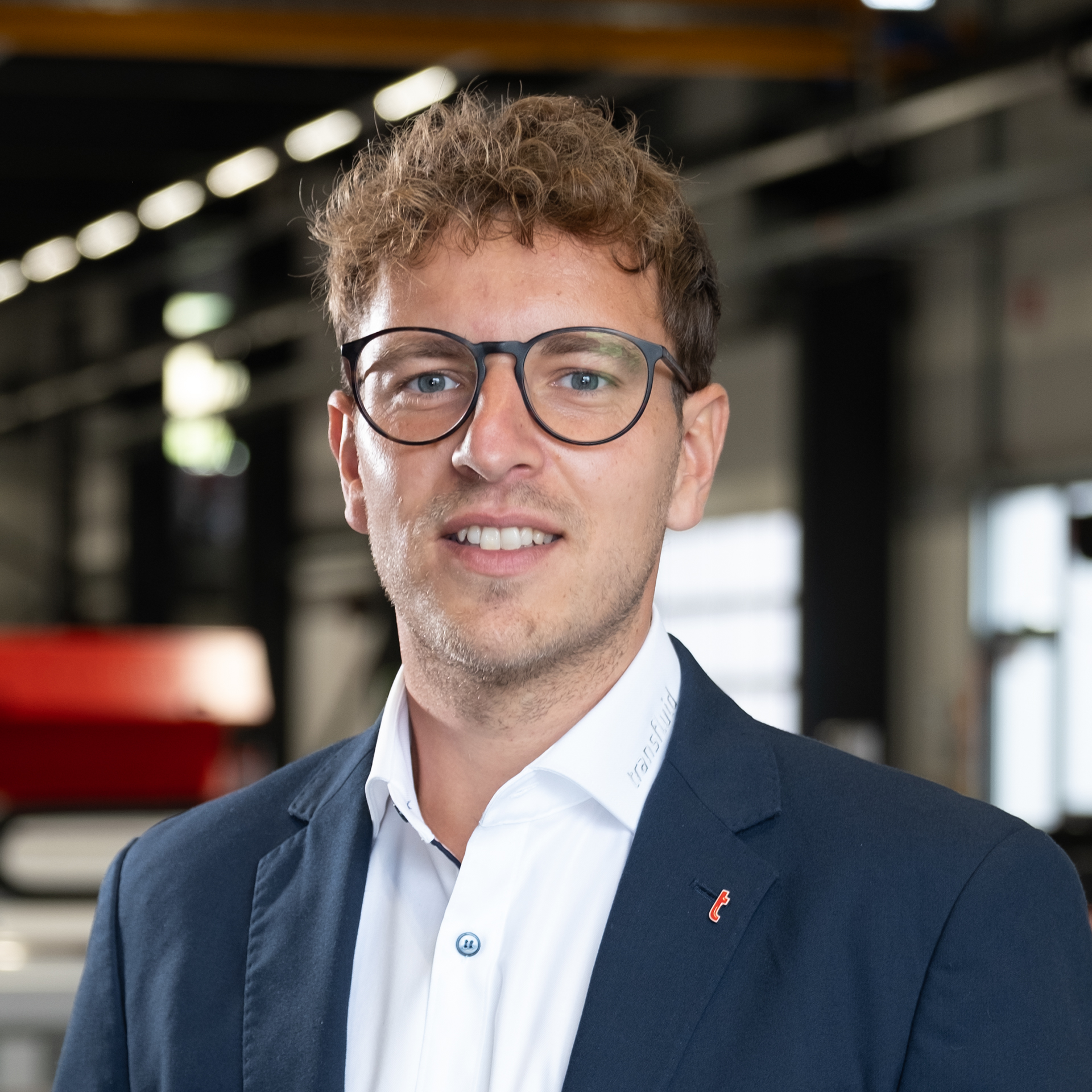 Mitarbeiter mit Brille im Anzug in Industriehalle mit Maschinen im Hintergrund.