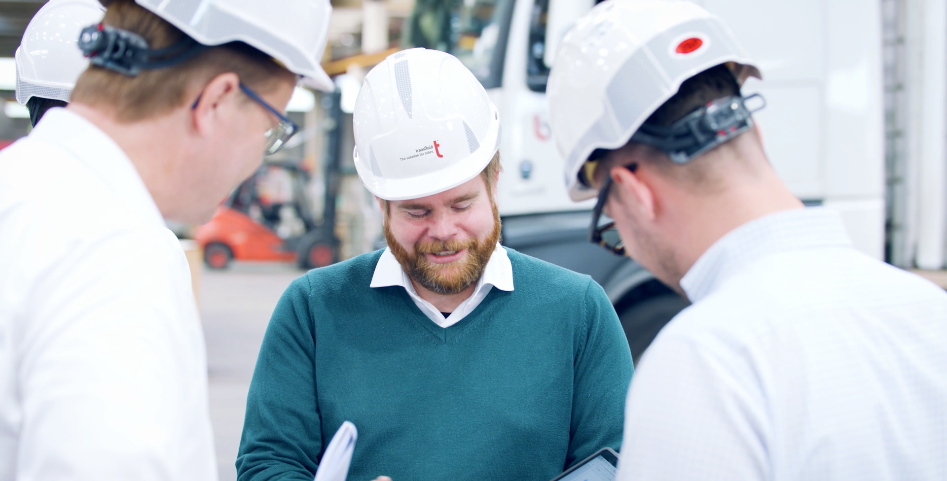 Drei Mitarbeiter mit Schutzhelmen besprechen Unterlagen in einer Industriehalle.