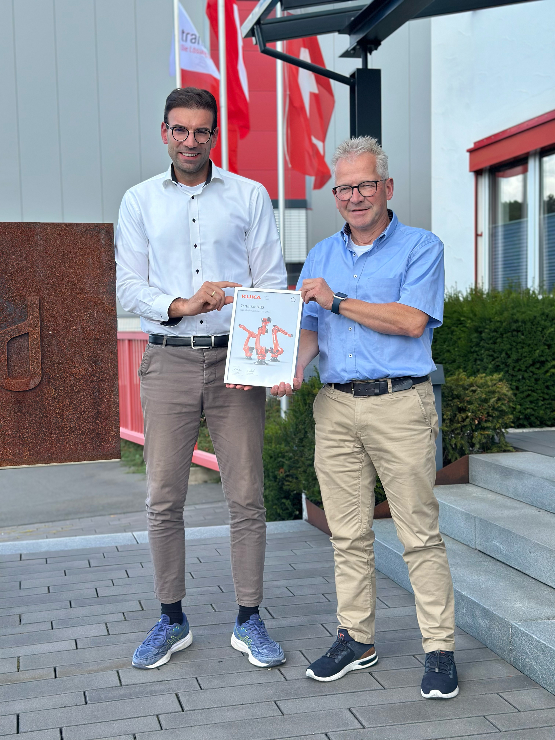 Zwei Männer präsentieren KUKA Partnerschaftszertifikat vor Firmengebäude.